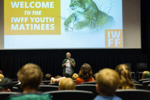 A person speaking to an audience in front of an IWFF slideshow.