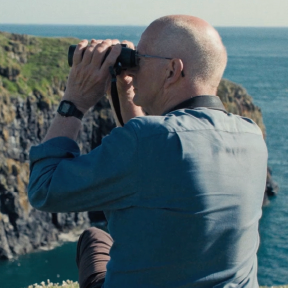 Person sits by cliffs and looks in binoculars.