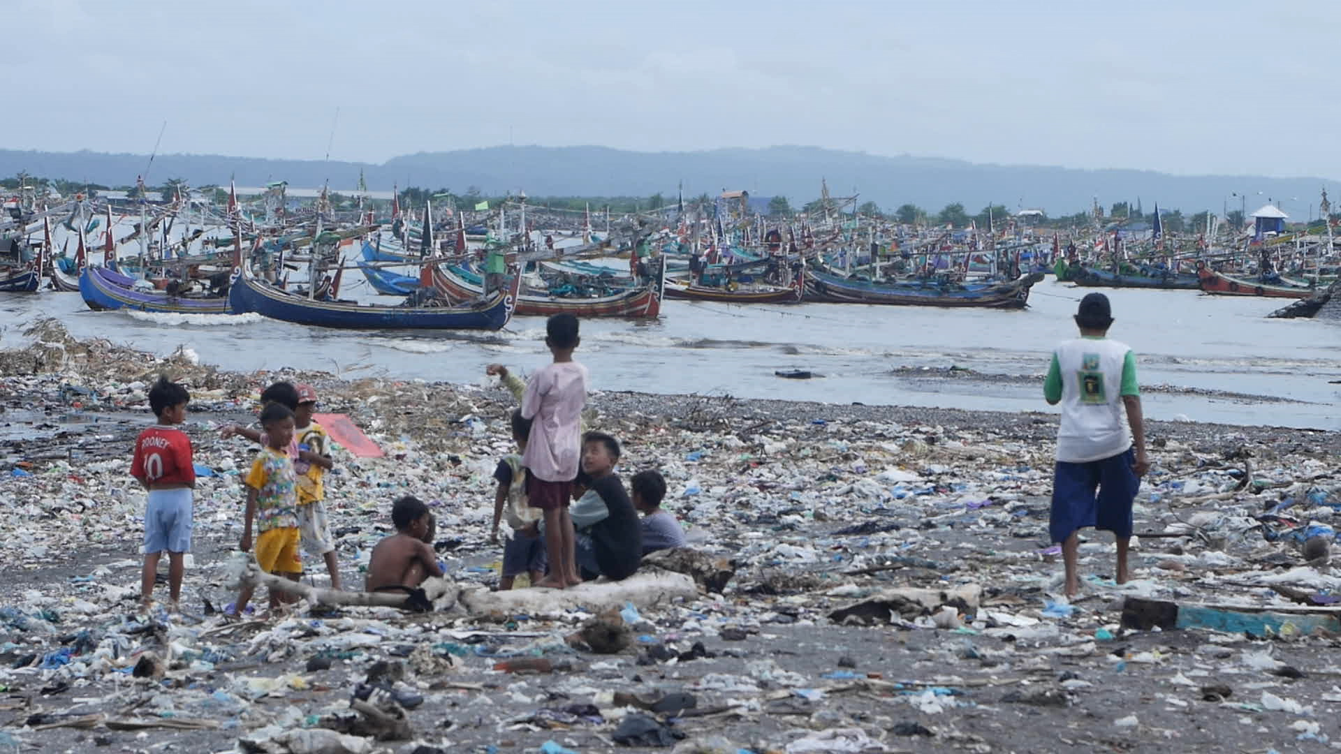 People stand on a beach full of trash looking at boats in water