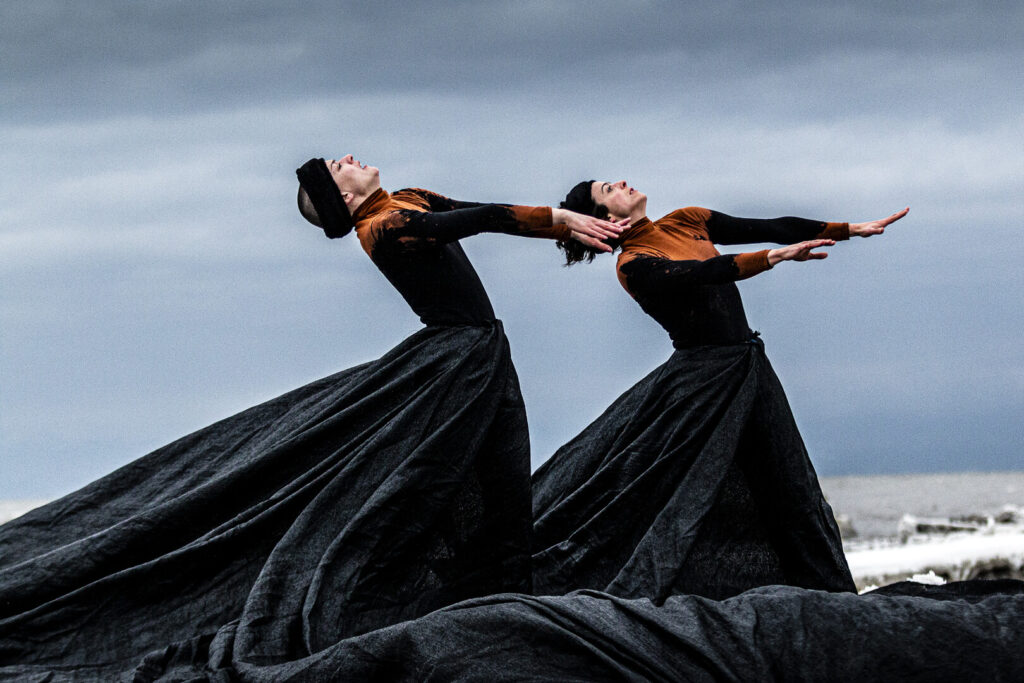 Two dancers in black with orange dresses moving outside.